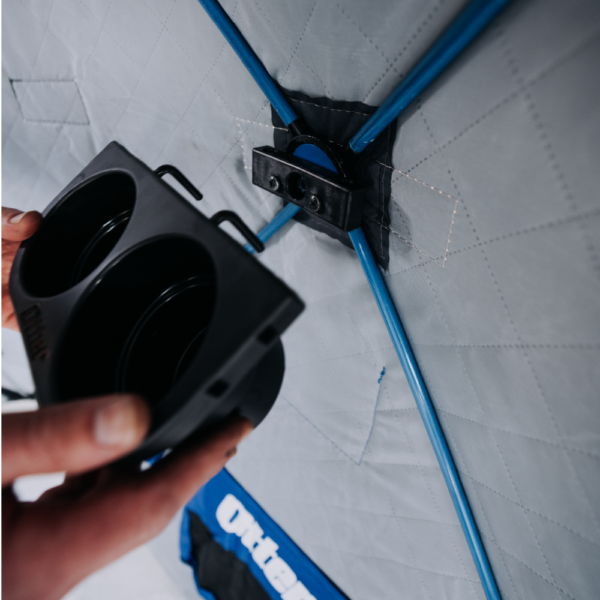Person's hands installing the black plastic Otter QA Dual Cup Holder onto the frame of a gray fabric fishing shelter with blue support rods.