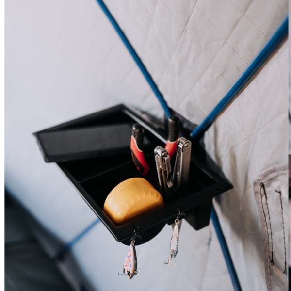 Otter QA Multi-Shelf mounted on an insulated tent wall, showing an open black plastic organizer containing tools and a yellow tape measure, with fishing lures hanging from hooks below.