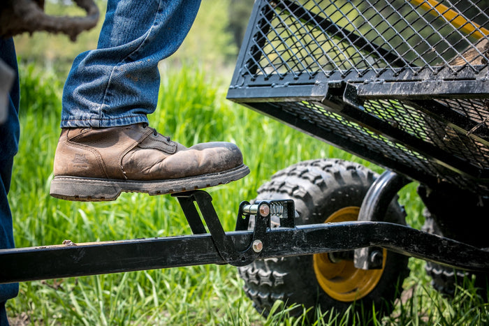 A person wearing brown leather boots presses down on a black metal lever attached to an outdoor utility cart wheel, demonstrating the Polar Foot Pedal Latch.