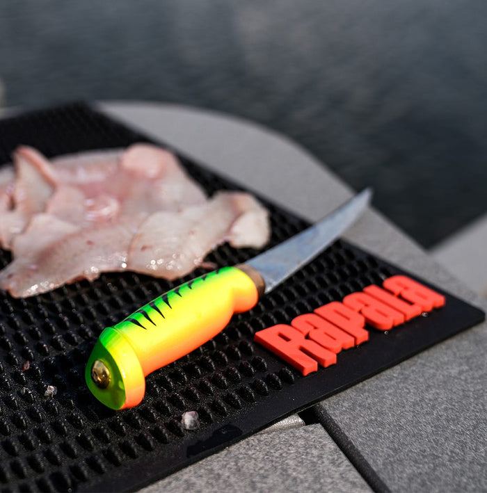 Rapala Fish 'N Fillet® Firetiger knife with a bright green and yellow handle beside fresh fish fillets on a textured cutting mat near water, designed for precise filleting.