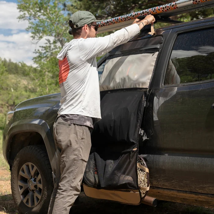 Man securing a patterned Riversmith Convoy Gear-Roll attached to the side of a dark vehicle, showing its multiple compartments and durable design for organized fishing tackle storage.