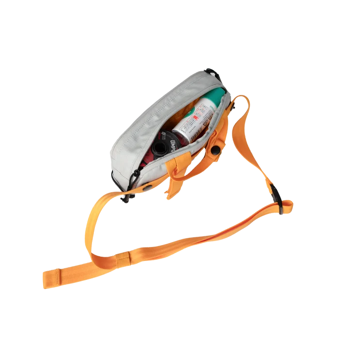 Riversmith Convoy Pack-Out Bag in gray with orange adjustable strap, shown open and partially filled with fishing tackle items and accessories for organized storage.