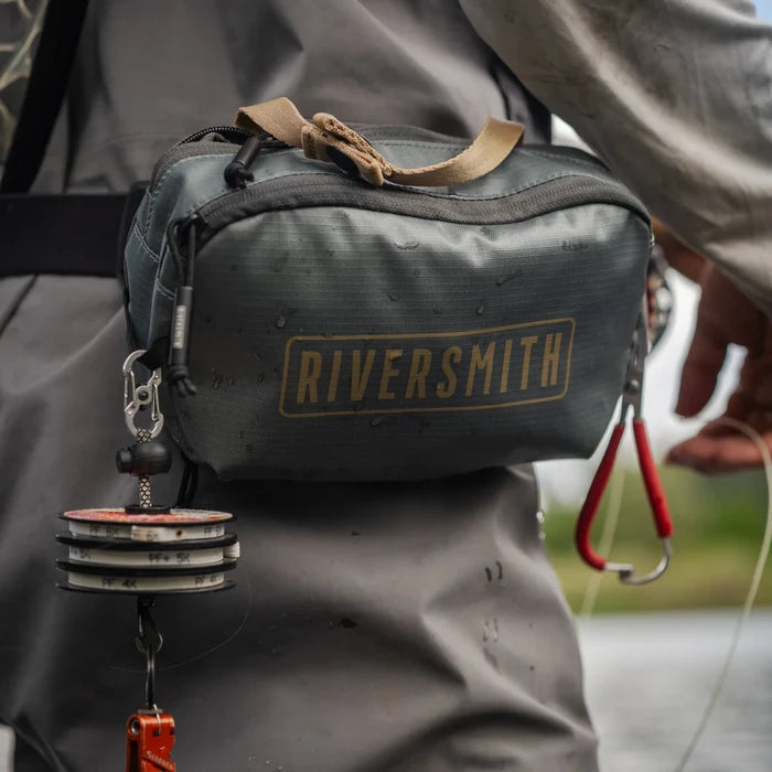 Riversmith Convoy Pack-Out Bag in gray worn around the waist with water droplets, featuring multiple compartments and attached fishing accessory tools for convenient tackle storage.