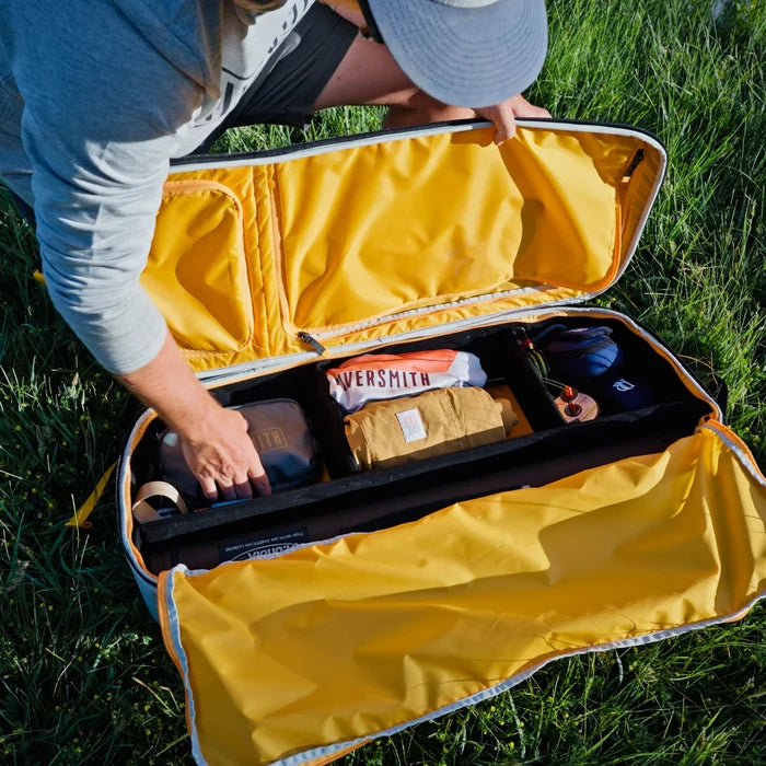 Person packing a bright yellow Riversmith Convoy Travel Pack with organized compartments holding fishing gear and outdoor essentials, shown open on grass.