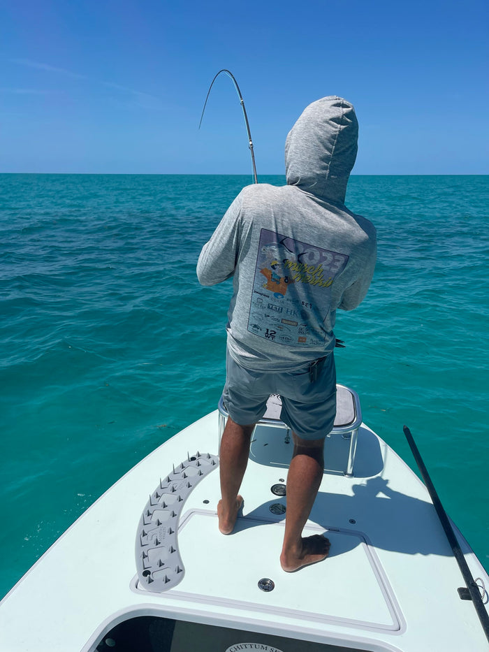 Angler standing barefoot on a boat casting a bent fishing rod wearing a hooded sweatshirt, showcasing the Scientific Anglers Launch Pad fishing rod against a clear blue ocean background.