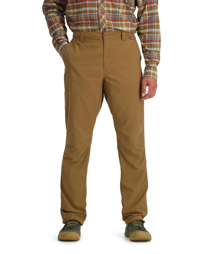 Man wearing tan Simms Bugstopper Superlight Pants paired with a plaid long-sleeve shirt and green outdoor shoes, standing with hands in pockets against a white background.