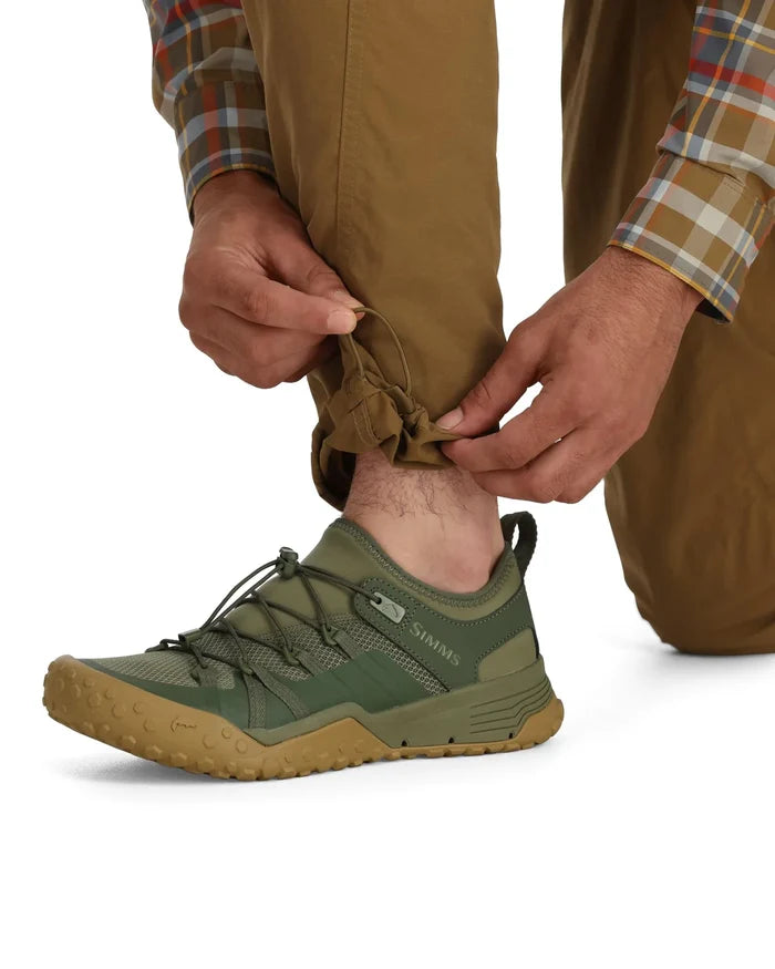 Close-up of a person adjusting the cuff of khaki Simms Bugstopper Superlight Pants above an olive green and tan fishing shoe, showing lightweight, durable fabric ideal for outdoor activities.