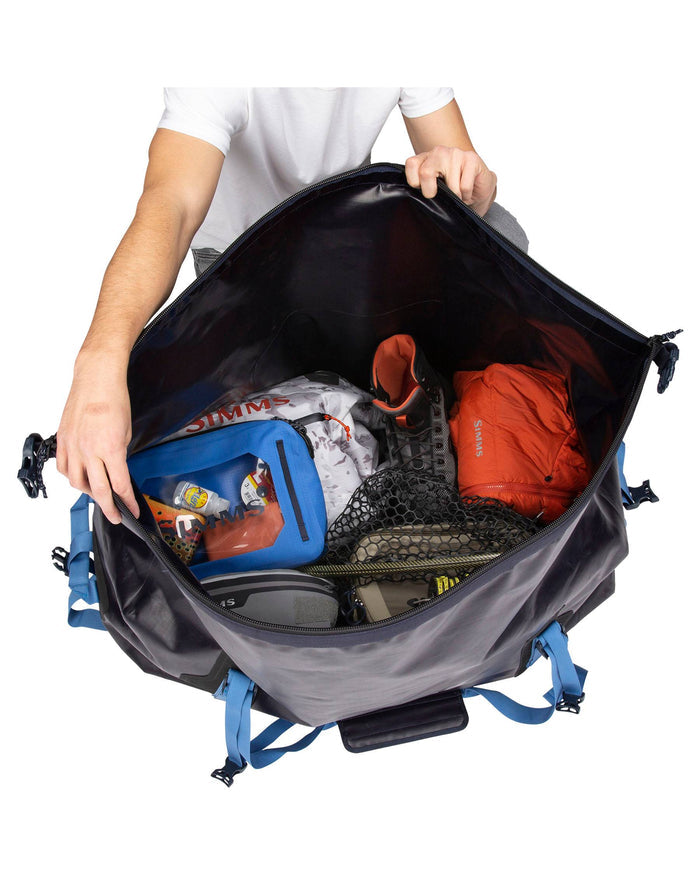 Simms Dry Creek Duffel opened to show a spacious interior packed with fishing gear including a waterproof blue pouch, fishing boots, net, and orange apparel, highlighting its storage capacity for tackle and outdoor equipment.
