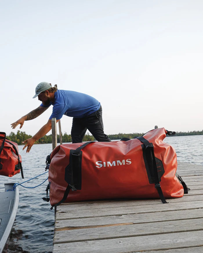 Simms Dry Creek Duffel in red, waterproof heavy-duty duffel bag with black straps, placed on a wooden dock next to a person reaching toward a boat by the water.