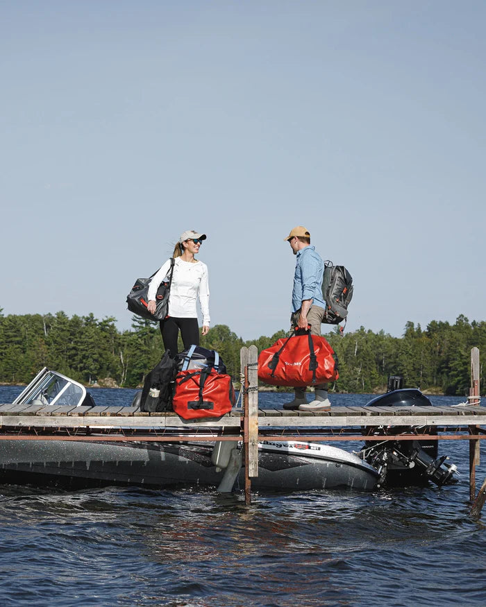 Two people standing on a wooden dock next to a boat, each carrying a gray backpack and holding a bright orange Simms Dry Creek Duffel bag, with a lake and forest in the background.