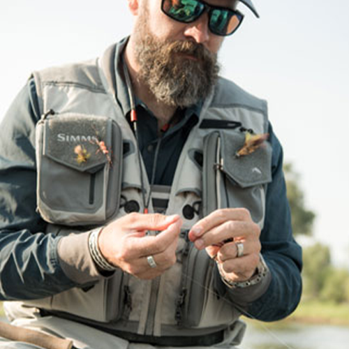 Man wearing a grey Simms G3 Guide Vest with multiple pockets and fishing gear, adjusting fishing line while seated outdoors near a blurred natural background.
