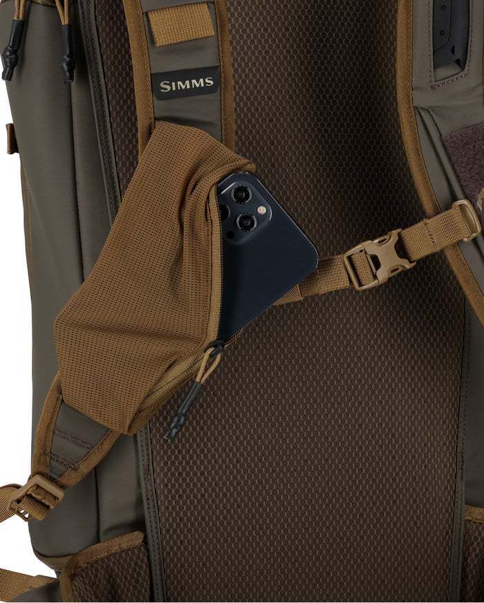 Simms Headwaters Backpack in brown with padded back panel and adjustable straps, showing a side zippered pocket holding a smartphone.