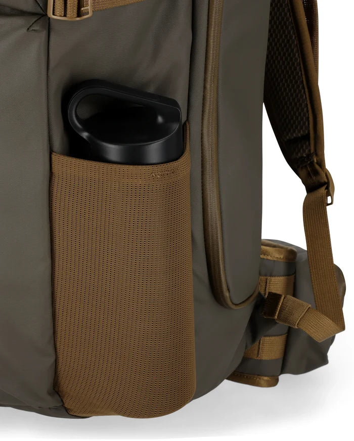 Close-up of the side pocket of a Simms Headwaters Backpack in olive green with brown mesh, holding a black water bottle, showing padded shoulder strap and durable fabric designed for fishing tackle storage.