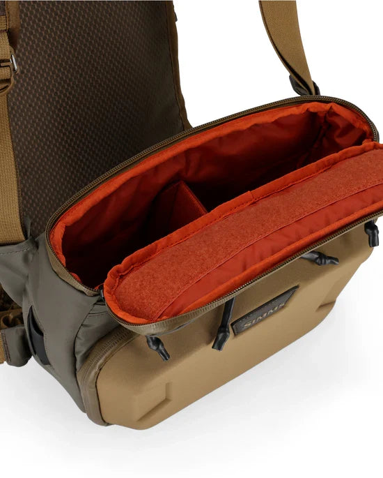Simms Headwaters Chest Pack in tan with an open top revealing a spacious main compartment lined with bright orange fabric and adjustable internal dividers for organized fishing tackle storage.