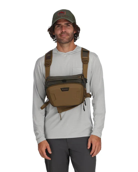 Man wearing a brown and olive Simms Headwaters Chest Pack with adjustable harness straps over a light gray long-sleeve shirt and dark pants, shown front view.