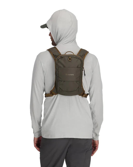 Man wearing a gray hooded shirt and dark pants shown from the back carrying an olive green Simms Headwaters Chest Pack with padded adjustable straps and multiple compartments for fishing tackle storage.