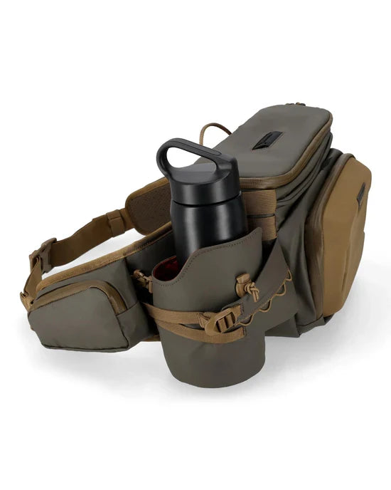 Simms Headwaters Hip Pack in muted green and brown with multiple zippered compartments and an insulated side pocket holding a black water bottle, designed for fishing tackle organization.