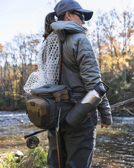 Angled view of a person wearing a gray jacket, cap, and sunglasses standing by a river with a Simms Headwaters Hip Pack holding fishing gear including a net, fishing rod, and water bottle.