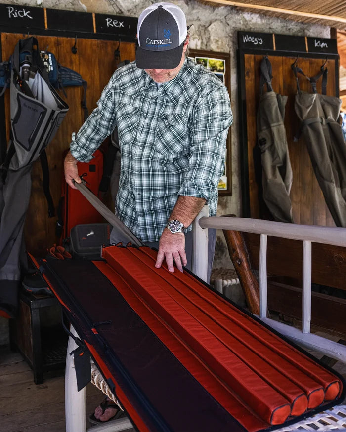 Man in plaid shirt and cap organizing red padded compartments inside a black Simms Riverkit Rod Roll on a white wooden bench in a rustic fishing gear shop.