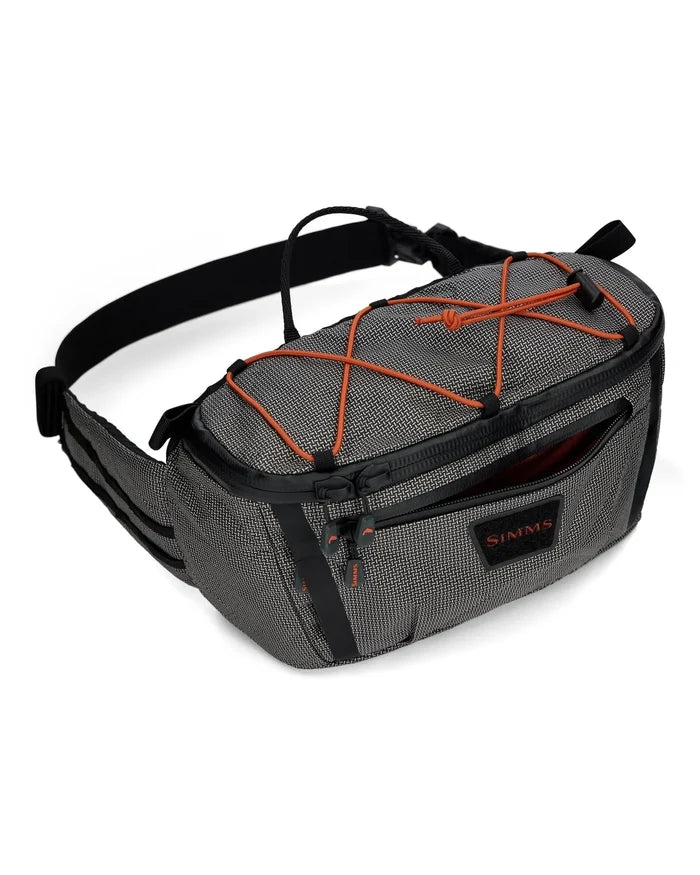 Simms Scout Hip Pack in gray fabric with black straps and orange bungee cord detailing, featuring zippered compartments and a compact design for fishing tackle storage.