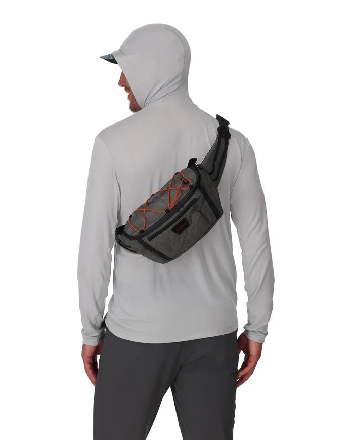 Man wearing a light gray hoodie and dark pants with a gray and black Simms Scout Hip Pack featuring orange bungee cord details worn across his back.
