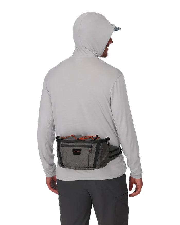 Man wearing a light gray hoodie and dark pants shown from the back, wearing a gray and black Simms Scout Hip Pack with orange accents around the waist, designed for fishing tackle storage and convenience.