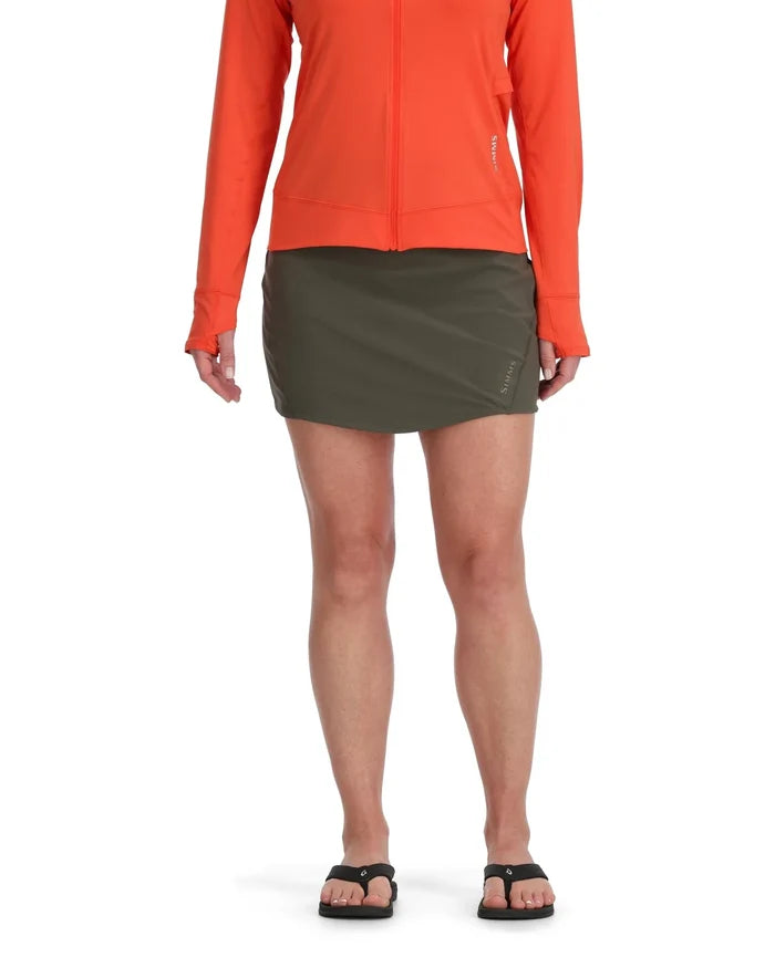 Simms Skort - Women's in olive green, worn with a bright orange zip-up jacket and black sandals, designed for comfort and mobility during fishing or outdoor activities.