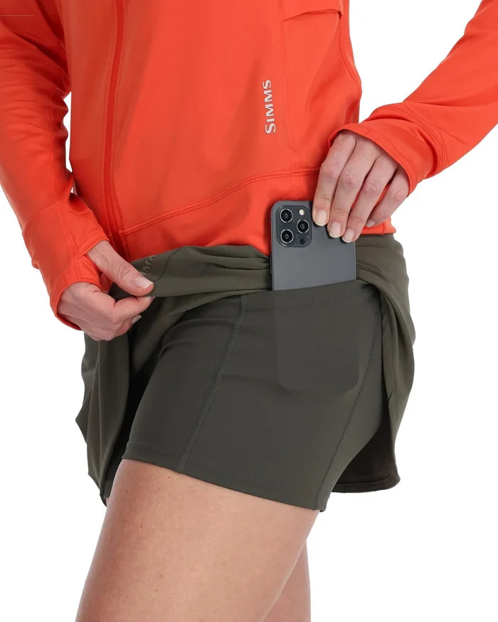 Close-up of a person wearing a green Simms Skort - Women's with built-in shorts, shown placing a smartphone into the side pocket, paired with an orange long-sleeve fishing shirt.