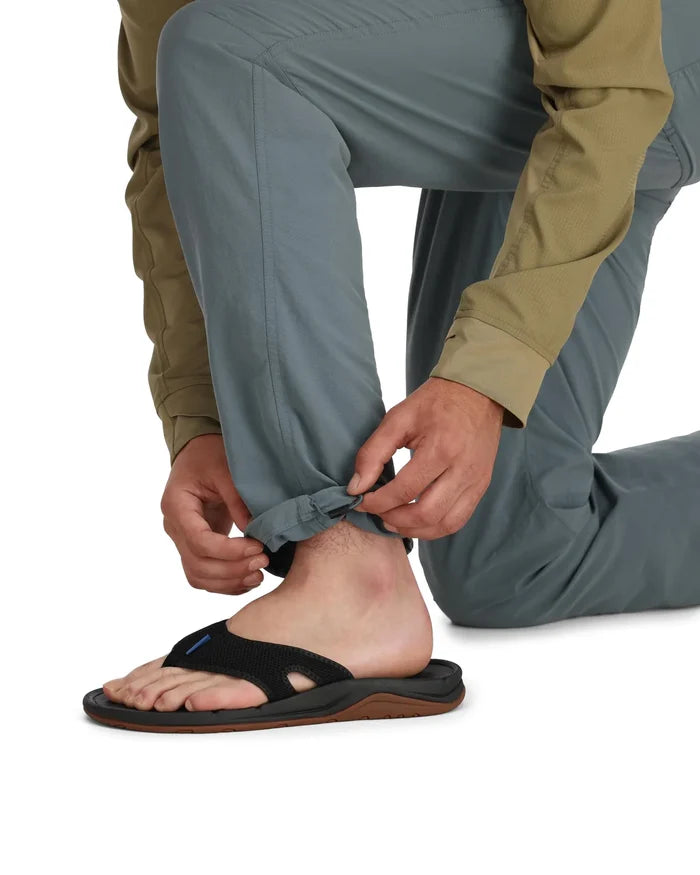 Man kneeling adjusts the cuff of gray Simms Superlight Pants (Previous Model) while wearing a tan long-sleeve shirt and black and brown flip-flops, showcasing lightweight fishing apparel.