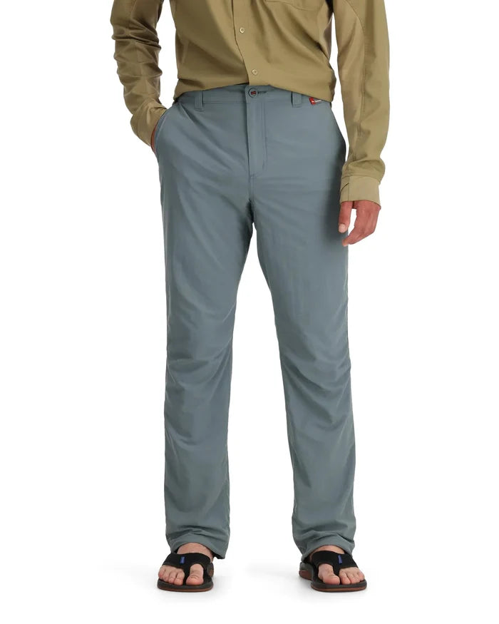 Man wearing light gray Simms Superlight Pants (Previous Model) with a tan long-sleeve shirt and black sandals, standing with hands in pockets against a white background.
