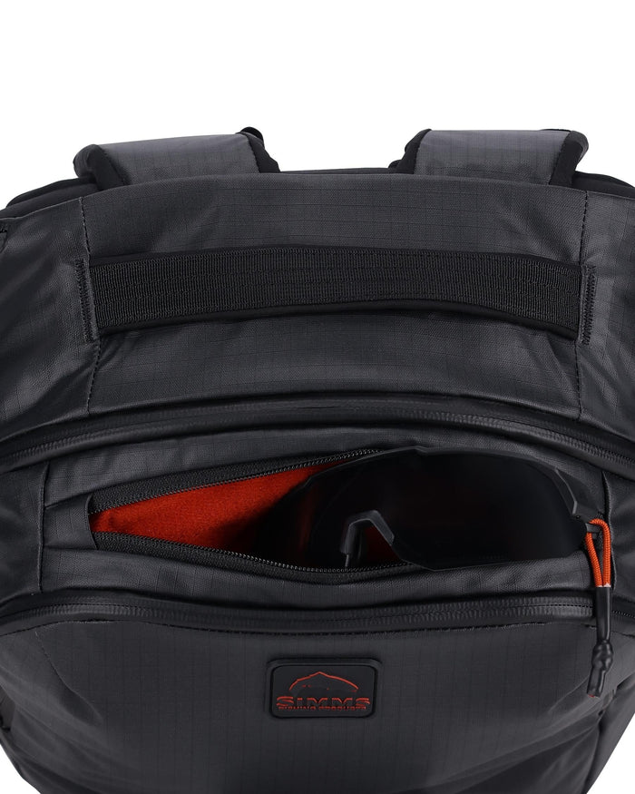 Close-up of the top section of a black Simms Tailwind Backpack showing a zippered compartment with an orange interior, designed for storing fishing tackle and accessories, featuring padded shoulder straps and a reinforced handle.