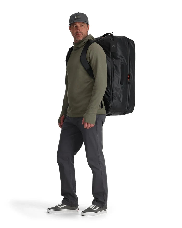 Man wearing casual outdoor clothing and a cap, carrying a large black Simms Tailwind Duffel 80L backpack-style bag with padded shoulder straps designed for fishing tackle and gear.