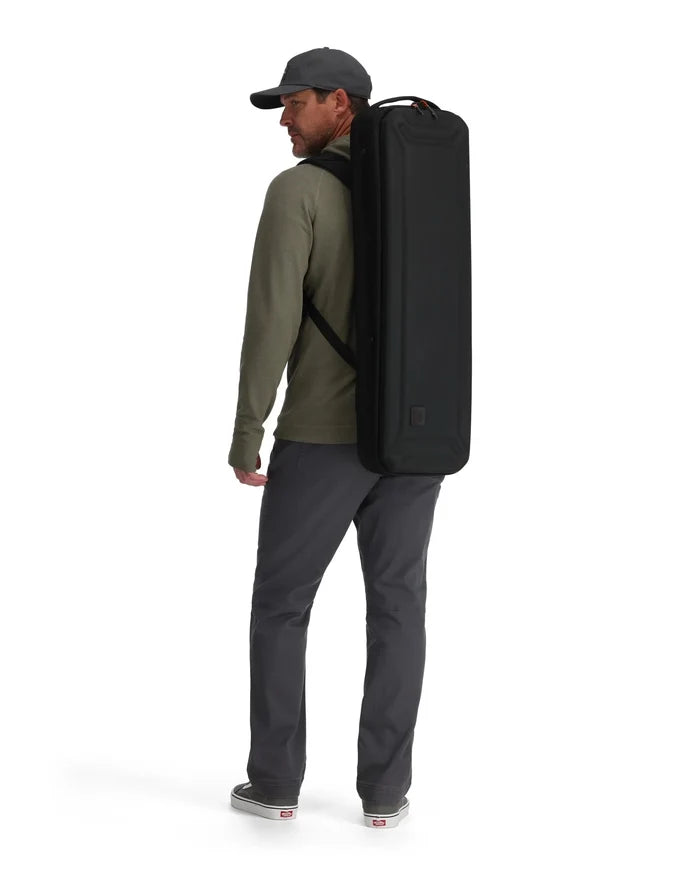 A man wearing a black cap and olive green long-sleeve shirt carries the Simms Tailwind Rod & Reel Vault on his back, a sleek black protective case designed for fishing rods and reels.