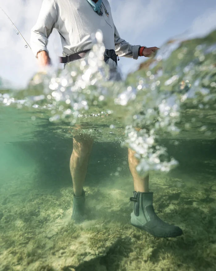 Person wearing water-resistant Simms Zip Bootie wading in clear shallow water above rocky riverbed, shown from below knee level with fishing rod visible.