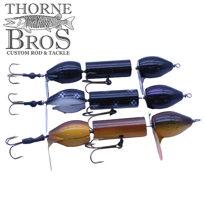 Three Snaggle Tooth Tackle Bar Bell fishing lures in black, gray, and brown finishes, each featuring dual treble hooks and a jointed body for realistic movement.