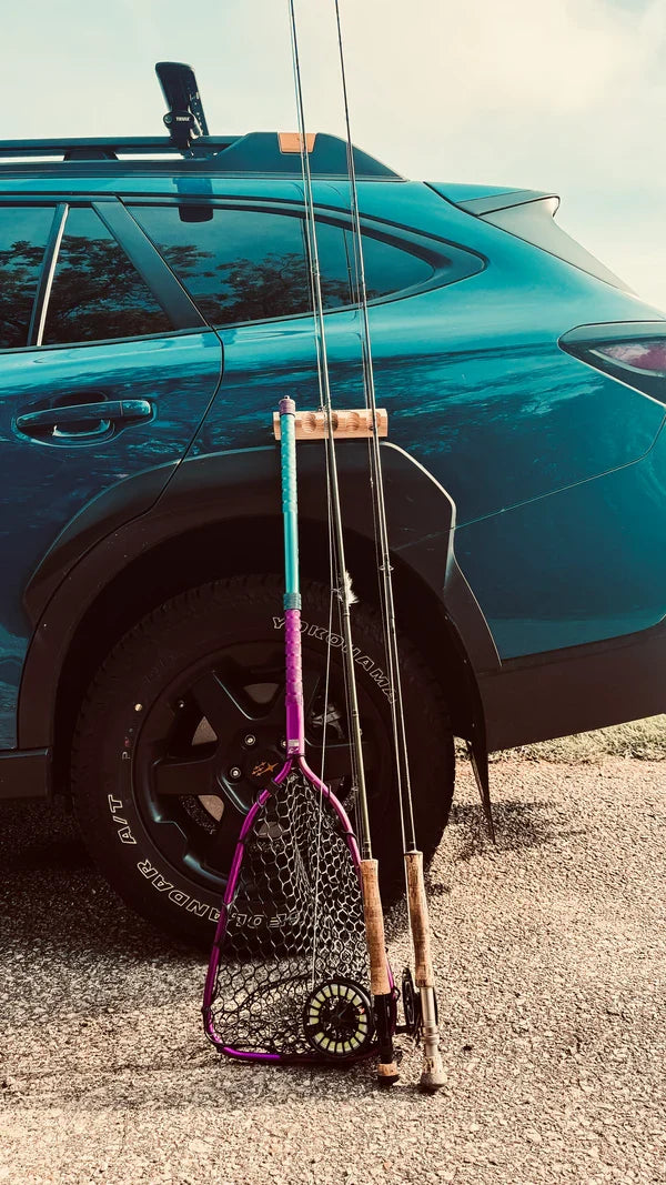 Temperance & Penn CastHitch-Magnetic Rod Holder attached to a blue SUV, securely holding two fishing rods and a landing net with a purple handle beside the rear tire on a gravel surface.