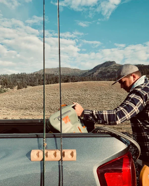 Man securing gear in the back of a pickup truck with two fishing rods held upright by a Temperance & Penn CastHitch-Magnetic Rod Holder mounted on the truck’s tailgate against a scenic outdoor backdrop.