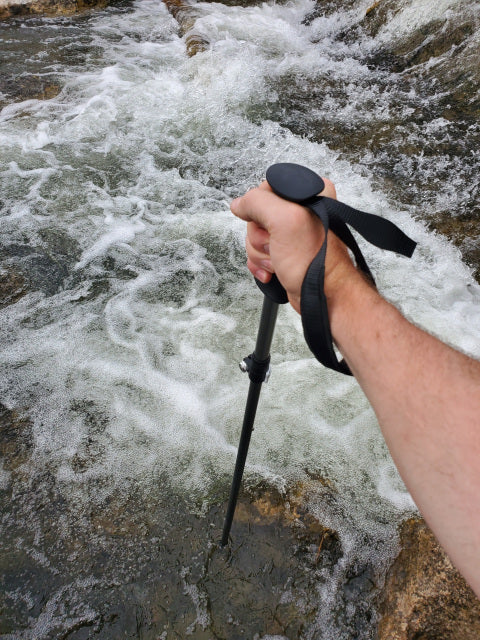 Person holding a Teton Locking Wading Staff with a black handle and wrist strap, stabilizing in a flowing rocky stream, designed for fishing tackle and outdoor support.