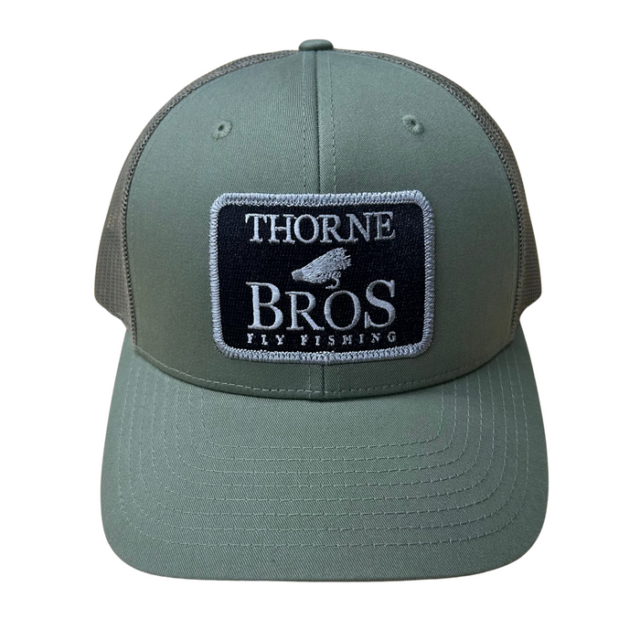 Thorne Bros Fly Fishing Logo Richardson 112 Trucker Cap in olive green with a black and white embroidered patch featuring a fly fishing lure design, mesh back, and curved brim.