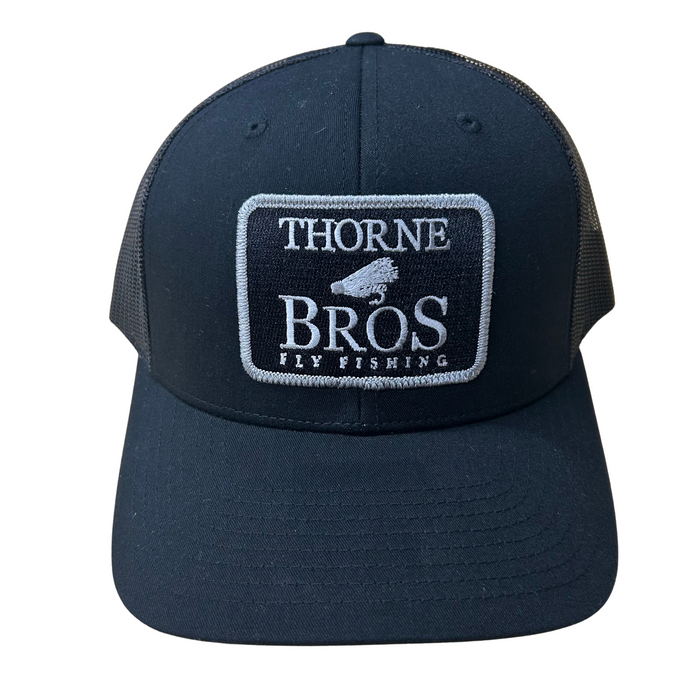 Thorne Bros Fly Fishing Logo Richardson 112 Trucker Cap in black with white embroidered patch featuring fishing lure icon and mesh back for breathability.