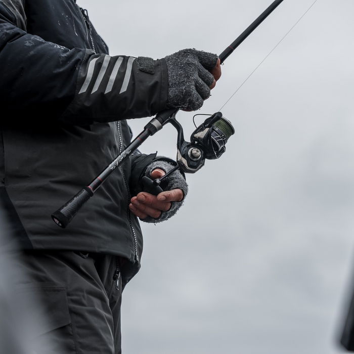 Angled close-up of a person wearing gloves and a black jacket holding a Thorne Bros Stealth Bass/Walleye Rods - Custom fishing rod with spinning reel against a cloudy sky.