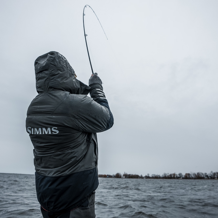 Angled view of an angler in a rain jacket casting a fishing rod over a large lake on a cloudy day, demonstrating the use of Thorne Bros Stealth Bass/Walleye Rods - Custom for freshwater fishing.