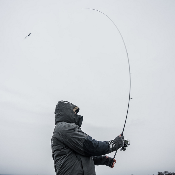 Angled view of an angler wearing a black hooded jacket casting a fishing lure using Thorne Bros Stealth Bass/Walleye Rods - Custom against an overcast sky background.