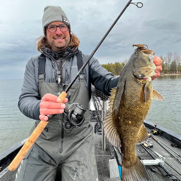 Man wearing gray hat and glasses holding a large bass fish in one hand and a Thorne Bros Stealth Bass/Walleye Rods - Custom fishing rod with cork handle and spinning reel in the other while sitting in a boat on a lake.