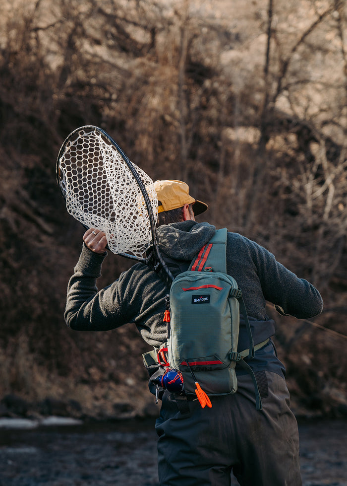 Angled rear view of a person fishing in a river wearing a green Umpqua Northfork Sling Pack with red accents, holding a large fishing net over their shoulder.