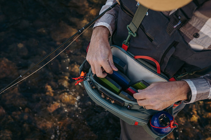 Angled top view of a person organizing fishing tools and tackle inside an open Umpqua Northfork Waist Pack while standing near clear water with a fishing rod beside them.
