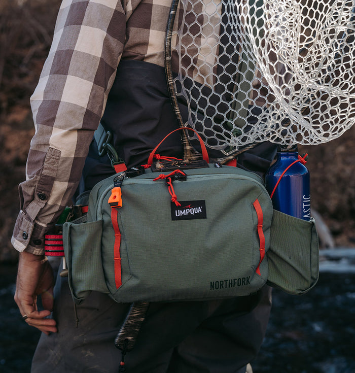 Umpqua Northfork Waist Pack in olive green with red accents worn by a person fishing, featuring multiple compartments, a fishing net, and a water bottle holders for organized tackle storage.
