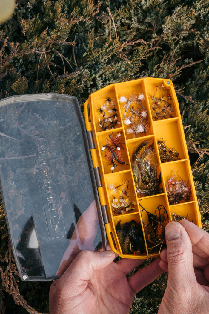 Umpqua UPG Compartment Fly Box in yellow with multiple compartments filled with various fishing flies, held open by a person's hands over natural outdoor foliage.