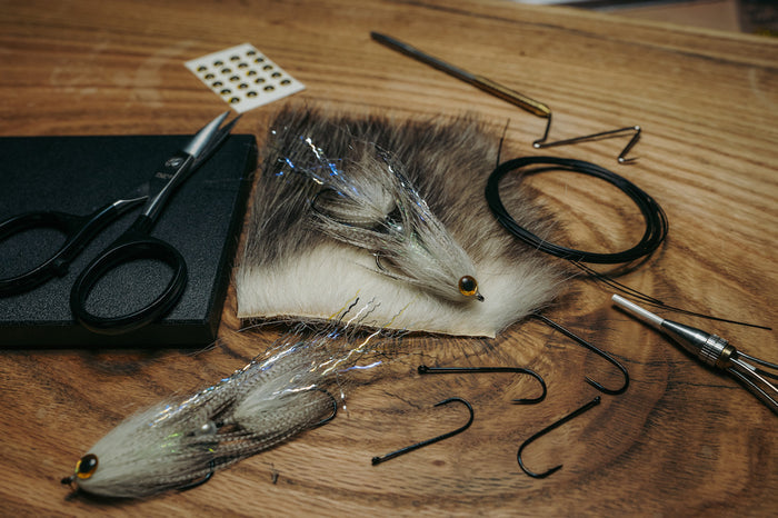 Umpqua X-Series XT350 fly fishing lures with iridescent fibers and realistic eyes, displayed alongside black fishing hooks, scissors, fur material, and fly tying tools on a wooden surface.
