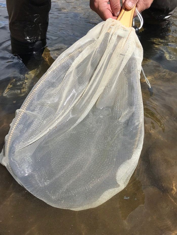 Hand holding a Wind River Gear Seine Nets / Entomology Kit with a fine mesh net and wooden handle, partially submerged in shallow water near fishing waders.